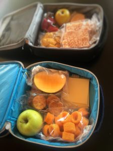 Two packed lunches in lunchboxes. Including fruit and processed foods.