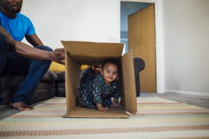 Mixed race baby girl crawling through a cardboard box in the sitting room at home, her father is holding the box up for her and her sister is in the background.